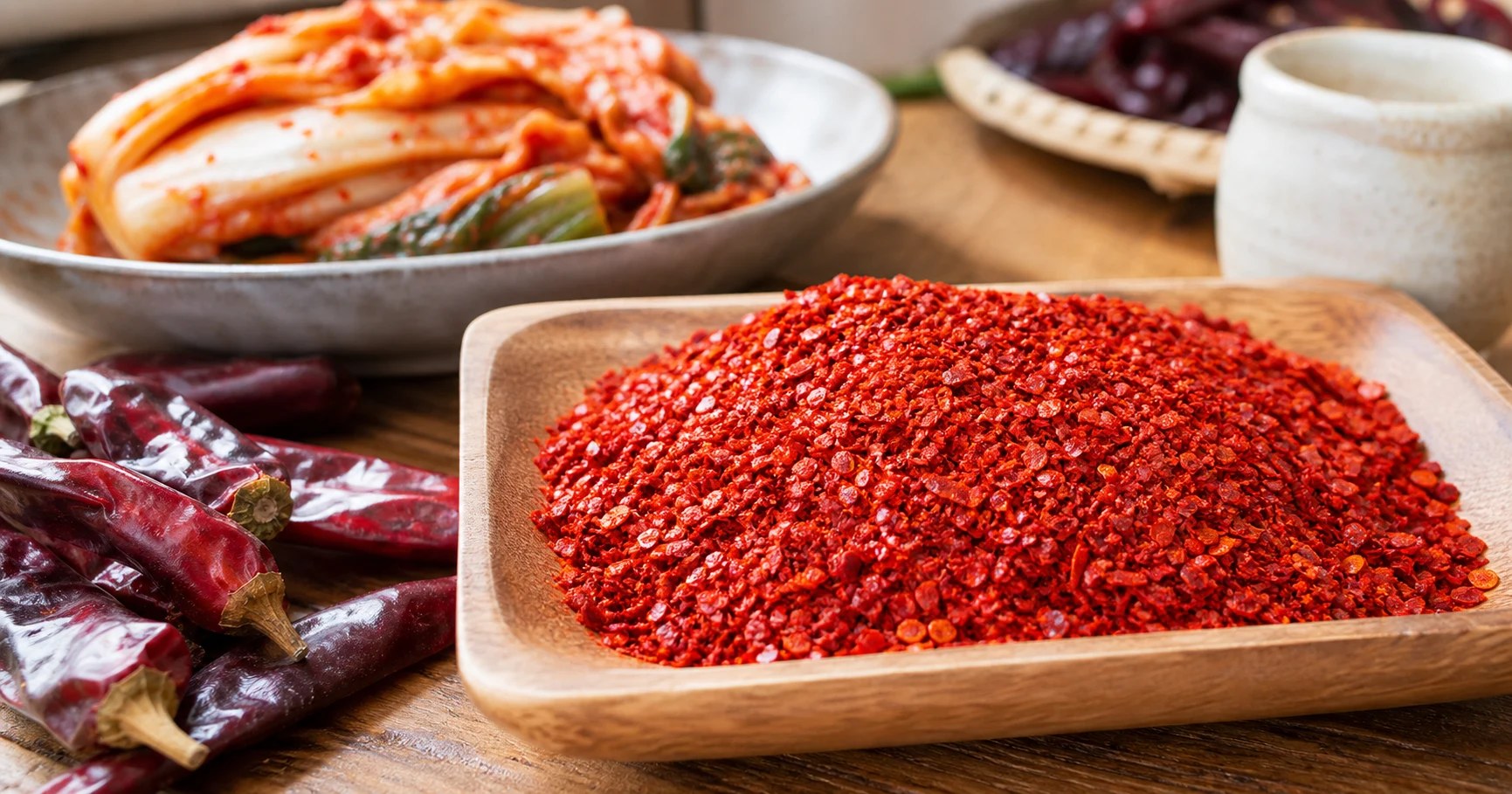 Coarse red gochugaru chili flakes beside dried Korean peppers and kimchi