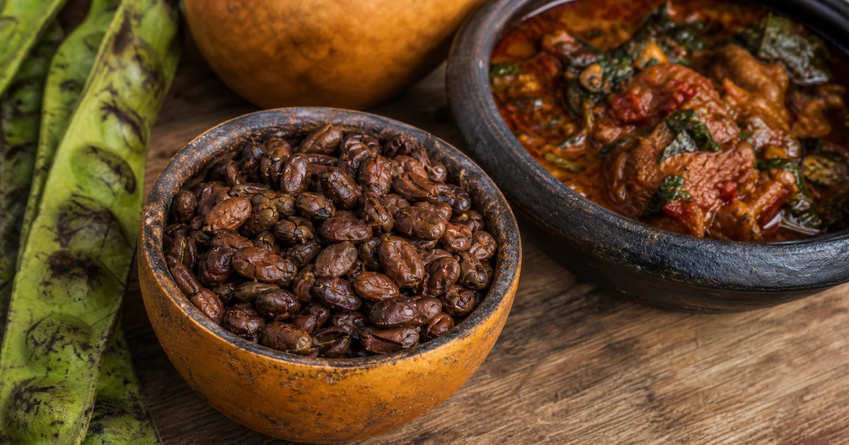 Brown fermented iru locust beans in a small calabash beside Parkia pods and stew