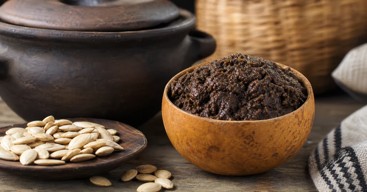 Dark West African ogiri fermented seed condiment beside melon seeds and a clay pot