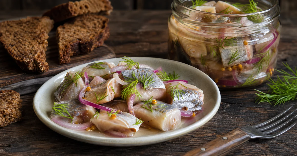 Pickled herring pieces with onion dill and spices in a glass jar beside rye bread