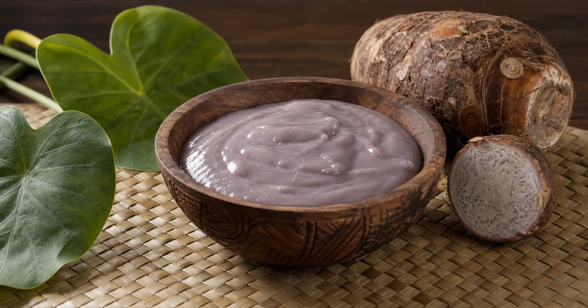 Purple-gray Hawaiian poi in a wooden bowl beside steamed taro corm and a stone pounder