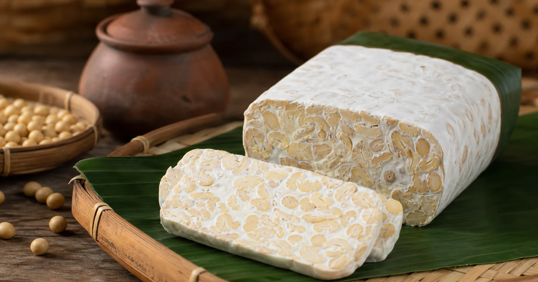 Sliced tempeh soybean cake wrapped in banana leaf beside a fermentation vessel